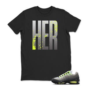 May include: Black t-shirt with the word "HER" in large, gray, block letters with neon green splatter accents. A pair of gray, black, and neon green athletic shoes are in the foreground.