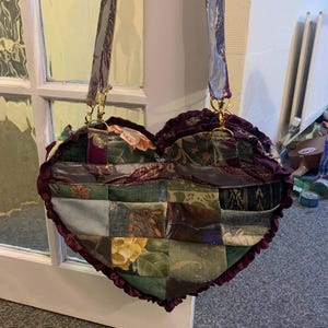 May include: A heart-shaped patchwork handbag with a ruffled edge and long straps. The bag features a variety of fabric patterns in shades of green, purple, and gold. The straps are a silvery-purple with gold hardware.