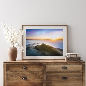 May include: A framed photograph of a hilltop with a stone tower at the summit, surrounded by fog and a colorful sunrise in the distance.
