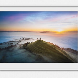 May include: A panoramic view of a green hilltop with a stone tower at the peak. The hill is covered in fog, with a bright orange sunrise in the background.
