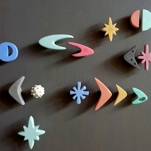 May include: A collection of 11 colorful ceramic knobs in various shapes, including stars, boomerangs, and circles. The knobs are arranged on a gray background.