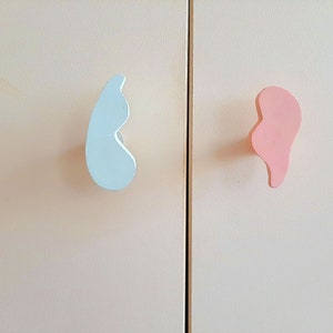 Op de afbeelding: Twee kastknoppen, één lichtblauw en één roze, zijn gevormd als abstracte organische vormen. De knoppen zijn gemonteerd op een witte kastdeur.