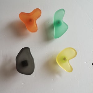 May include: Four colorful wall hooks in the shape of abstract forms. The hooks are made of translucent plastic and have a metallic finish. The hooks are in orange, green, gray, and yellow.