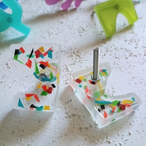 Peut inclure: Deux boutons de cabinet en verre transparent avec un motif de confettis colorés. Les boutons ont une vis en métal argenté et une boule en verre transparent sur le dessus.