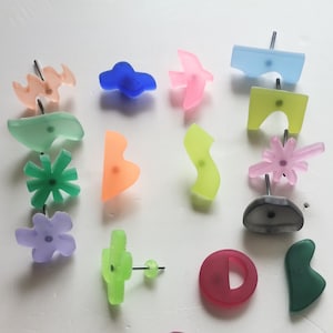 Puede incluir: Surtido de alfileres de empuje de resina de formas abstractas y geométricas de colores. Los alfileres están sobre una superficie blanca e incluyen formas como una flor, una estrella, un pájaro, un corazón y un círculo.