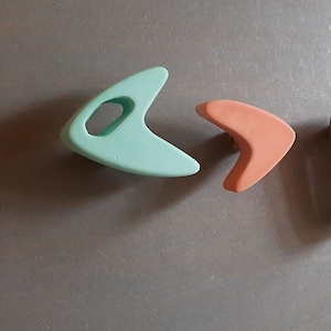 May include: A light mint green boomerang-shaped object with a central oval cutout and a smaller, solid peach-colored boomerang-shaped object. Both items feature smooth, matte surfaces and curved, pointed ends, presented as distinct decorative pieces.