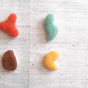 May include: Four handmade felt shapes in vibrant colors: orange, teal, brown, and yellow. The shapes are organic and abstract, arranged against a white brick background. These could be decorative elements or craft supplies.