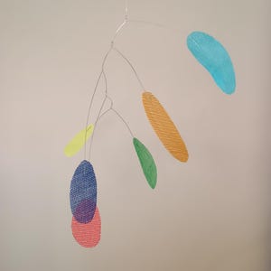 Puede incluir: Móvil colorido con elementos en forma de hojas en varios colores, incluyendo azul, naranja, verde y rosa. Las hojas están suspendidas de finos alambres plateados, creando una obra de arte abstracta y moderna. El móvil está diseñado para colgar y girar.