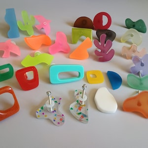 May include: Assortment of colourful, abstract, and geometric shaped cabinet knobs and drawer pulls. The knobs are made of acrylic and have a variety of colours, including pink, blue, green, yellow, orange, red, and purple.