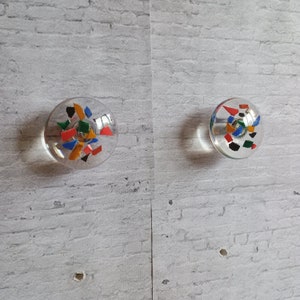 Peut inclure: Deux boutons en verre transparent avec des motifs géométriques colorés. Les boutons sont ronds et ont une base en métal argenté.