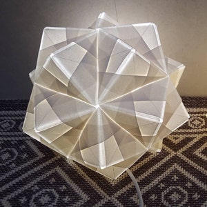 May include: A white origami lampshade with a geometric design. The lampshade is made of paper and has a delicate, intricate pattern.