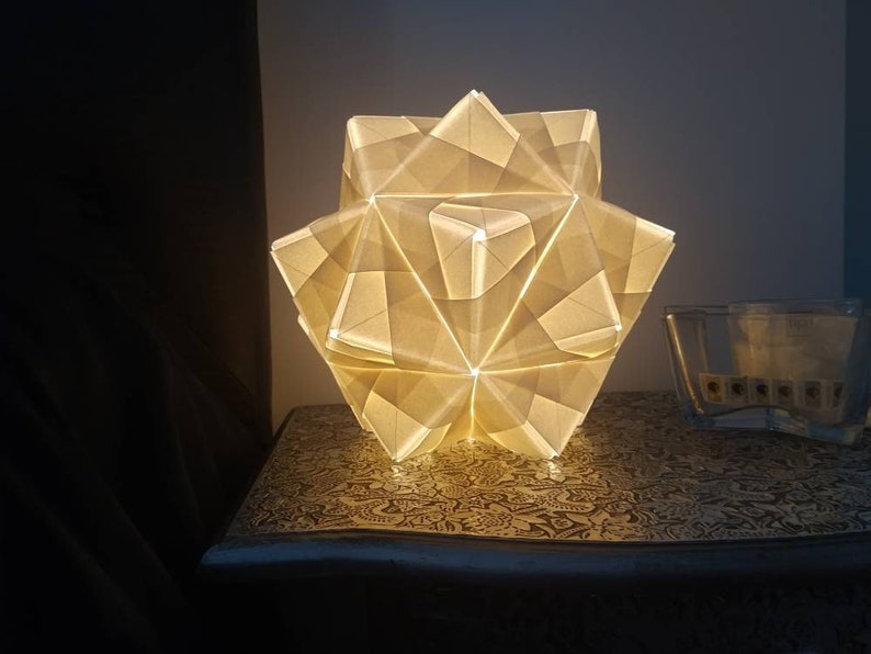 May include: A white origami lamp with a star-shaped design, illuminated from within, sits on a patterned table.