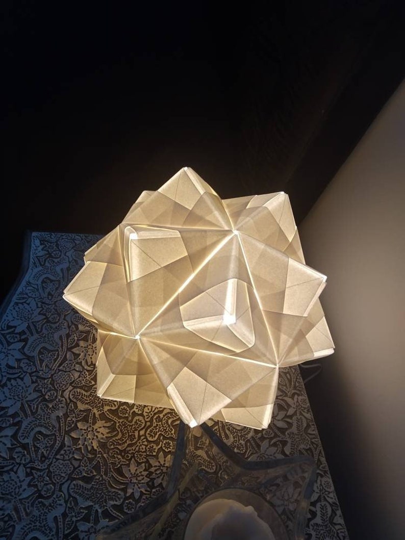 May include: A white origami lampshade with a geometric design. The lampshade is lit from within, casting a warm glow.