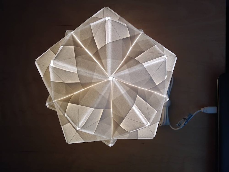 May include: A white origami-style lamp with a geometric design. The lamp is lit and casts a warm glow.