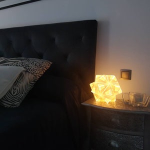 May include: A white geometric lamp with a warm glow sits on a silver nightstand next to a bed with a black headboard and a zebra patterned pillow.