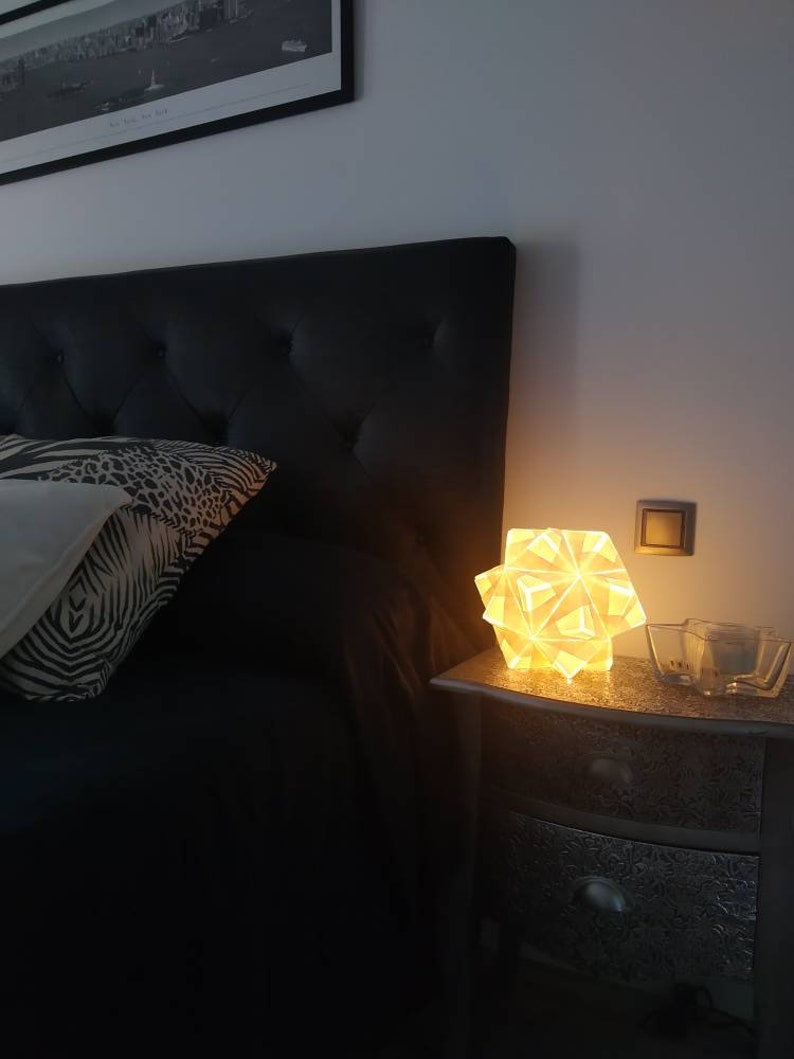 May include: A white geometric lamp with a warm glow sits on a silver nightstand next to a bed with a black headboard and a zebra patterned pillow.