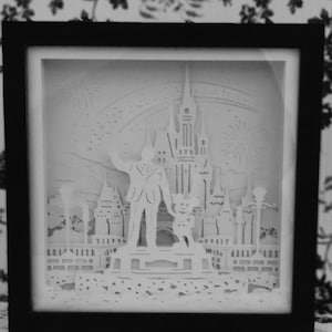 Könnte beinhalten: Eine weiße Papier-Schattenbox mit einer 3D-Silhouette von Schloss Aschenputtel, Mickey Mouse und einem Mann im Anzug. Das Schloss ist von Feuerwerk und einem Regenbogen umgeben.