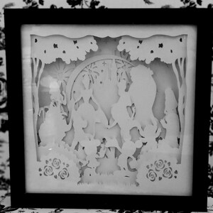 Könnte beinhalten: Ein weißes Papier-Schnittbild in einem schwarzen Rahmen. Das Kunstwerk zeigt eine Szene mit drei Figuren, einem Kaninchen und Blumen. Die Figuren stehen in einer Waldkulisse mit einem Halbmond im Hintergrund.