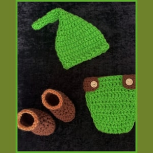 Puede incluir: Sombrero, cubrepañal y botines de crochet verde para un bebé. El sombrero tiene una punta puntiaguda y el cubrepañal tiene dos botones marrones. Los botines son marrones con ribete verde.