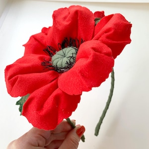 May include: A large, vibrant red felt poppy flower with a green and black center and a green stem. The handmade floral decoration has detailed petals and is a striking craft item.