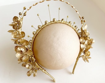 Könnte beinhalten: Ein goldfarbenes Kopfstück mit floralen und perlenbesetzten Akzenten. Das Kopfstück hat ein kreisförmiges Design mit einem zentralen beigefarbenen Kissen und ist mit zarten goldenen Blumen und kleinen Perlen verziert. Das Gesamtdesign ist elegant und aufwendig.