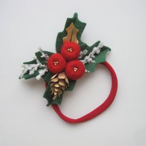 May include: A red elastic headband featuring a decorative arrangement of three red felt berries, green felt holly leaves, a small gold pinecone, a gold leaf accent, and white glittered sprigs that resemble frosted branches for a festive appearance.