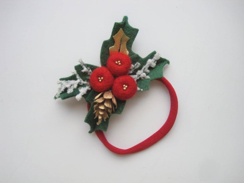 May include: A red elastic headband featuring a decorative arrangement of three red felt berries, green felt holly leaves, a small gold pinecone, a gold leaf accent, and white glittered sprigs that resemble frosted branches for a festive appearance.