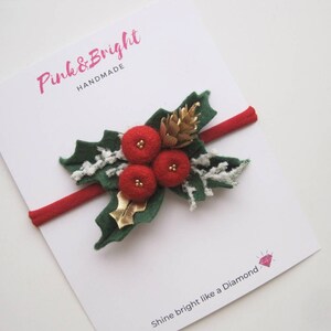 May include: A red elastic headband featuring a felt holly decoration with three red berries, green leaves, a gold pinecone, and white frosted accents. The display card features the text Pink & Bright HANDMADE and Shine bright like a Diamond.