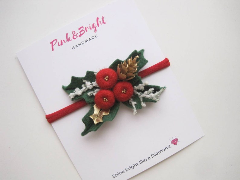 May include: A red elastic headband featuring a felt holly decoration with three red berries, green leaves, a gold pinecone, and white frosted accents. The display card features the text Pink & Bright HANDMADE and Shine bright like a Diamond.