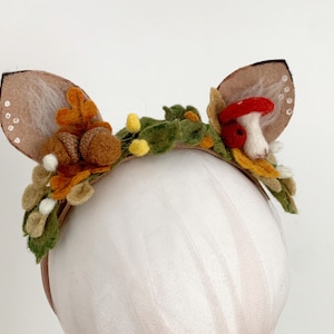 May include: A felt headband with deer ears, a red mushroom, acorns, and leaves. The headband is brown and the leaves are green, orange, and yellow.