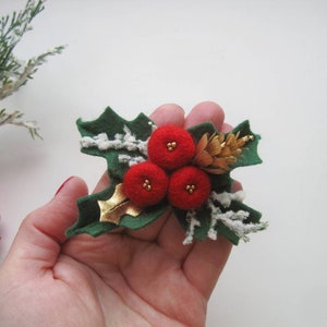 May include: A decorative brooch featuring three round red felt berries with small gold bead centers, dark green felt holly leaves, a small brown pinecone, a metallic gold leaf accent, and white textured snowy detailing on the green leaves.