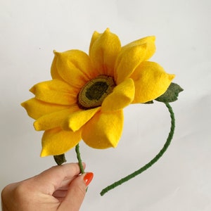 May include: A headband featuring a large, bright yellow felt sunflower with pointed petals and a textured, dark olive green center. The band itself is wrapped in a thin, green fabric, and a small green felt leaf is attached near the flower base.
