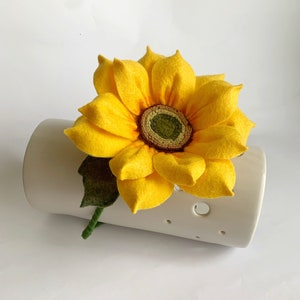 May include: A handcrafted felt sunflower with bright yellow petals and a textured green and brown center. The flower features a single dark green felt leaf and a wrapped green stem, resting against a cylindrical white ceramic object with holes.