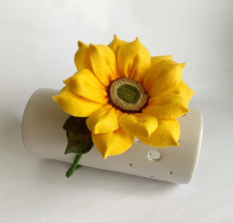 May include: A handcrafted felt sunflower with bright yellow petals and a textured green and brown center. The flower features a single dark green felt leaf and a wrapped green stem, resting against a cylindrical white ceramic object with holes.