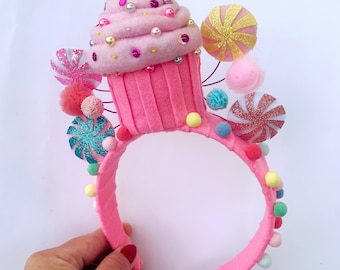 Könnte beinhalten: Ein pinkfarbenes Filz-Stirnband mit einem großen Cupcake und Süßigkeiten-Dekorationen. Der Cupcake hat einen pinkfarbenen Zuckerguss-Wirbel mit Perlen- und Pailletten-Akzenten. Bunte Pompons und glitzernde Lollis schmücken das Stirnband, was ein skurriles, festliches Accessoire ergibt.