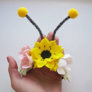Op de afbeelding: Een haarband met een grote gele viltzonnebloem, kleinere roze en witte viltbloemen en twee zwarte pijpenrager-antennes met gele pompons aan de uiteinden. Het bloemstuk is gecentreerd op een donkere band met groene viltblaadjes.