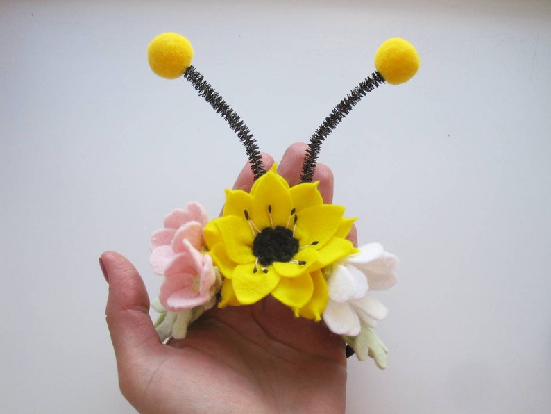 Op de afbeelding: Een haarband met een grote gele viltzonnebloem, kleinere roze en witte viltbloemen en twee zwarte pijpenrager-antennes met gele pompons aan de uiteinden. Het bloemstuk is gecentreerd op een donkere band met groene viltblaadjes.