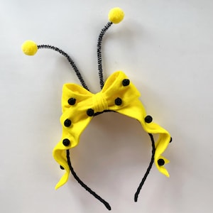 May include: A black headband featuring a bright yellow fabric bow with small black polka dots. Two black pipe cleaner antennae extend upwards from the centre, each topped with a small yellow pom-pom, creating a bee-inspired design.