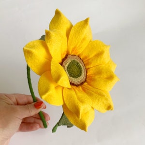 May include: A handcrafted felt sunflower featuring bright yellow petals arranged in multiple layers around a textured olive green center. The flower is attached to a thin, green-wrapped stem with a single green felt leaf extending from the base.