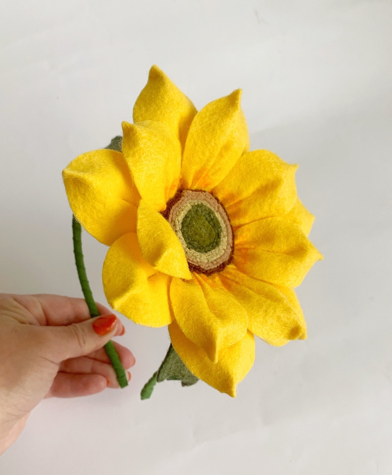 May include: A handcrafted felt sunflower featuring bright yellow petals arranged in multiple layers around a textured olive green center. The flower is attached to a thin, green-wrapped stem with a single green felt leaf extending from the base.