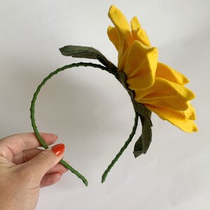 May include: A headband featuring a large, bright yellow felt flower with multiple layered petals. The headband frame is wrapped in a textured green material, and dark green felt leaves are attached at the base of the flower, extending outward.