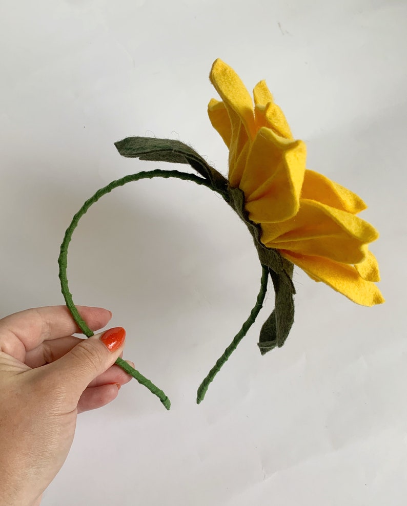 May include: A headband featuring a large, bright yellow felt flower with multiple layered petals. The headband frame is wrapped in a textured green material, and dark green felt leaves are attached at the base of the flower, extending outward.