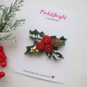 May include: A felt holly brooch featuring three red pom-pom berries, a gold metallic leaf, and a small gold pinecone on green felt leaves with white faux snow accents. The brooch is attached to a white card labeled Pink & Bright HANDMADE.