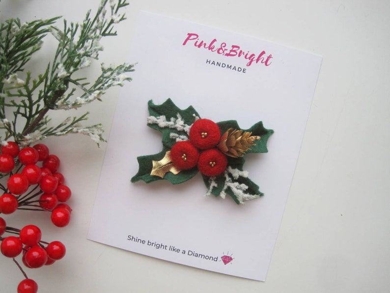 May include: A felt holly brooch featuring three red pom-pom berries, a gold metallic leaf, and a small gold pinecone on green felt leaves with white faux snow accents. The brooch is attached to a white card labeled Pink & Bright HANDMADE.