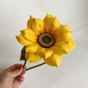 May include: A handcrafted felt sunflower featuring bright yellow petals with orange shading at the base. The center is a textured dark green disc surrounded by a ring of light green. A green felt leaf and a thin green stem are attached to the flower.
