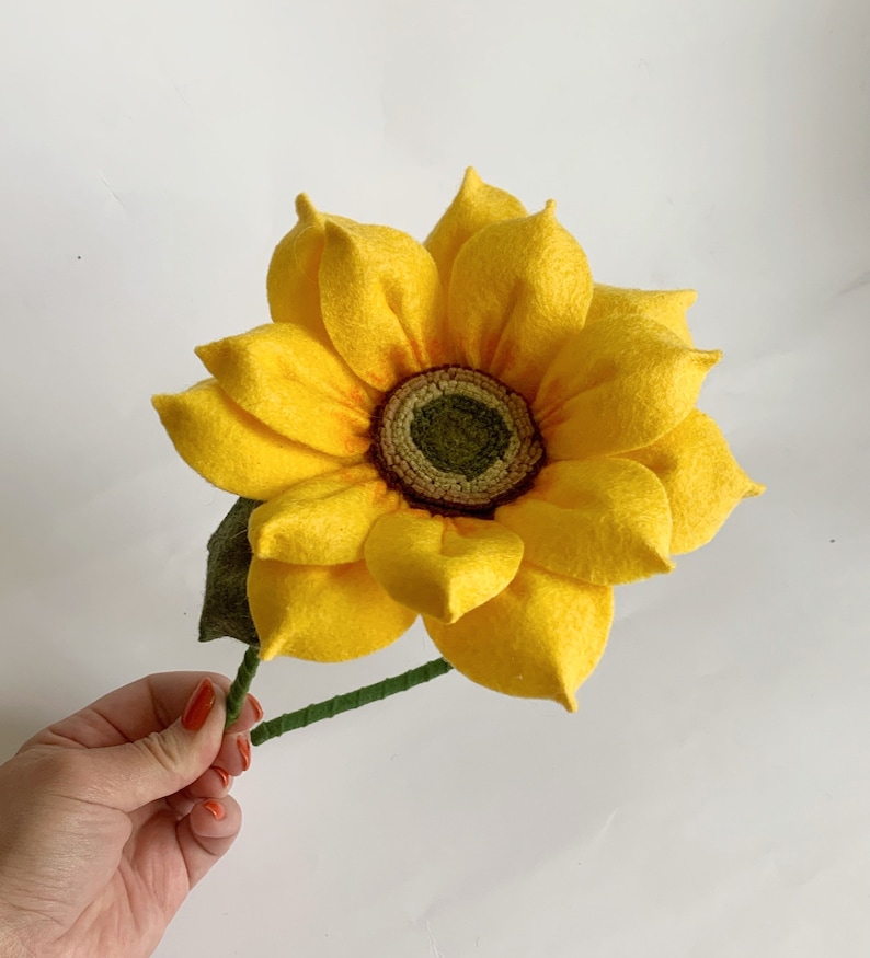 May include: A handcrafted felt sunflower featuring bright yellow petals with orange shading at the base. The center is a textured dark green disc surrounded by a ring of light green. A green felt leaf and a thin green stem are attached to the flower.