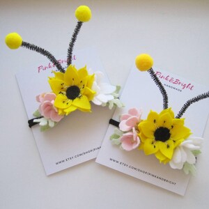 Op de afbeelding: Twee haarbanden met bijenthema, voorzien van viltbloemen in geel, wit en roze. Elke haarband heeft twee zwarte pijpenrager-antennes met gele pompons. De haarbanden worden gepresenteerd op witte kaarten met Pink & Bright Handmade erop.