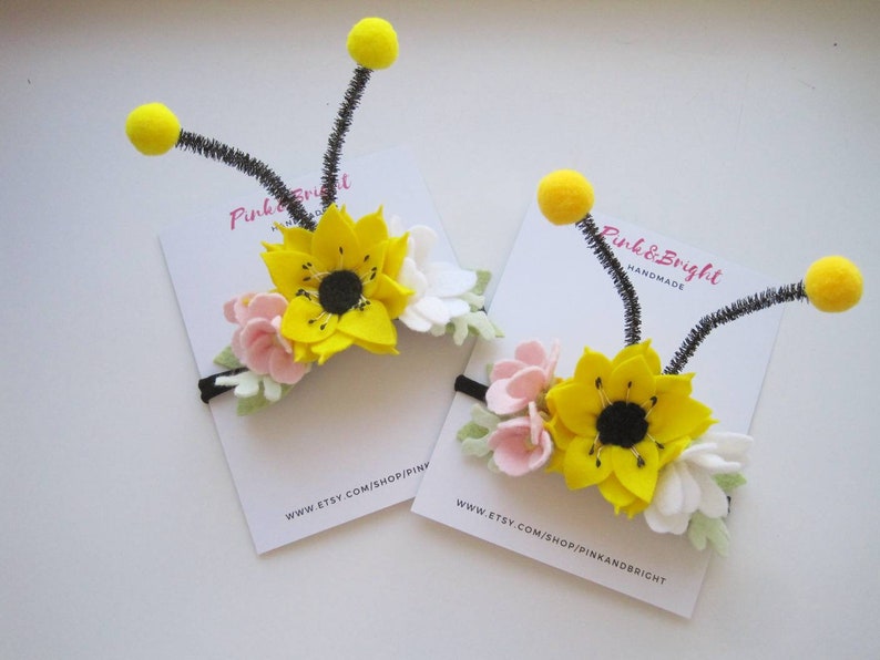 Op de afbeelding: Twee haarbanden met bijenthema, voorzien van viltbloemen in geel, wit en roze. Elke haarband heeft twee zwarte pijpenrager-antennes met gele pompons. De haarbanden worden gepresenteerd op witte kaarten met Pink & Bright Handmade erop.