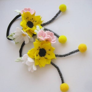 Op de afbeelding: Twee zwarte haarelastiekjes versierd met viltbloemen in geel, wit en lichtroze. Elk haarelastiekje heeft een grote gele bloem met een zwart hart, kleinere witte en roze bloemetjes en twee zwarte pijpenragers met gele pompons aan de uiteinden.