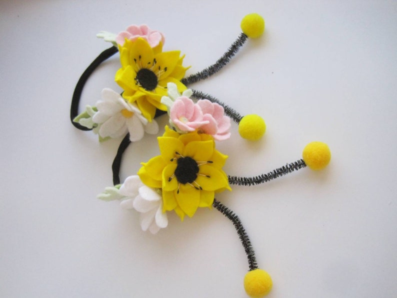 Op de afbeelding: Twee zwarte haarelastiekjes versierd met viltbloemen in geel, wit en lichtroze. Elk haarelastiekje heeft een grote gele bloem met een zwart hart, kleinere witte en roze bloemetjes en twee zwarte pijpenragers met gele pompons aan de uiteinden.